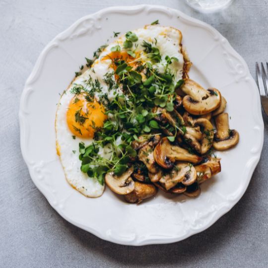 A white plate with eggs, mushrooms, potatoes, and microgreens.