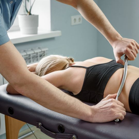 Caucasian male chiropractor performing Instrument Assisted Soft Tissue Mobilization on a caucasian female patient.