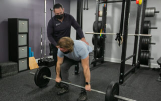 Male chiropractor cueing male patient on proper bracing during a deadlift.