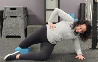 Female chiropractor demonstrating a modified Copenhagen plank using a foam block for pelvic stability and SPD support in a chiropractic clinic.