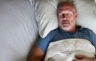 Middle-aged man sleeping on his back in bed, head on a pillow and covered with a blanket.