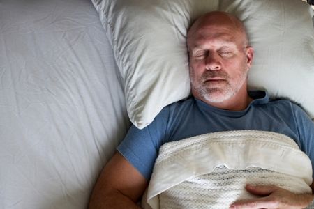 Middle-aged man sleeping on his back in bed, head on a pillow and covered with a blanket.